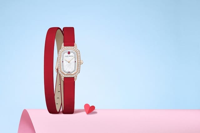 Elegant red leather wristwatch with a diamond-studded face on a pink and blue background with a small red heart accent.