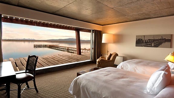 Hotel room with two beds, large window view of a serene lake and pier, cozy chairs, and soft lighting for a relaxing ambiance.