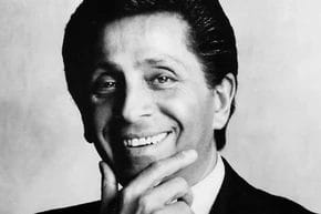 Man smiling, wearing a suit, with hand on chin, black and white photo.