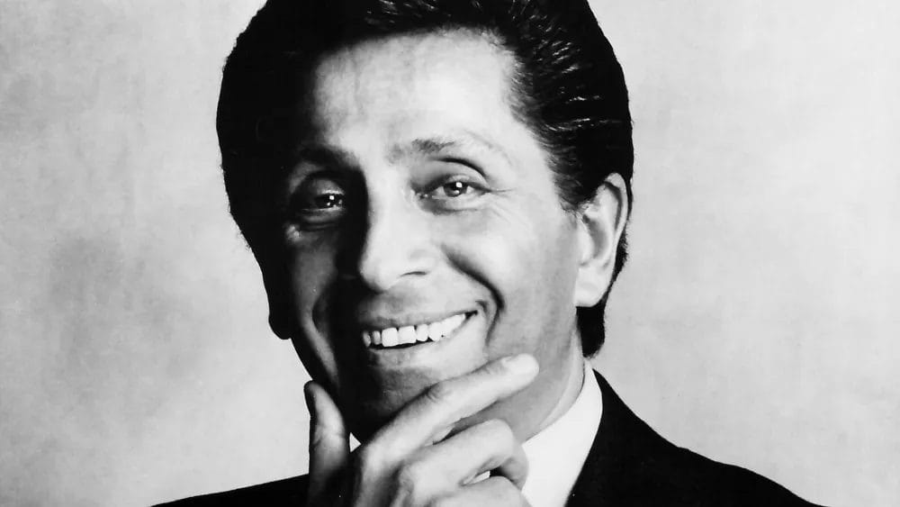 Man smiling, wearing a suit, with hand on chin, black and white photo.