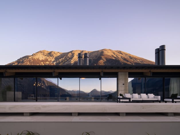Modern house with large glass windows and mountain view at sunset, featuring outdoor seating area on a spacious patio.