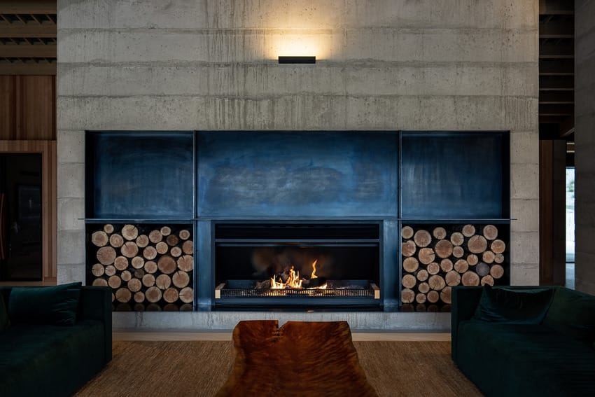 Modern fireplace with burning logs, concrete wall, and wood storage, flanked by dark green sofas in a cozy living room.