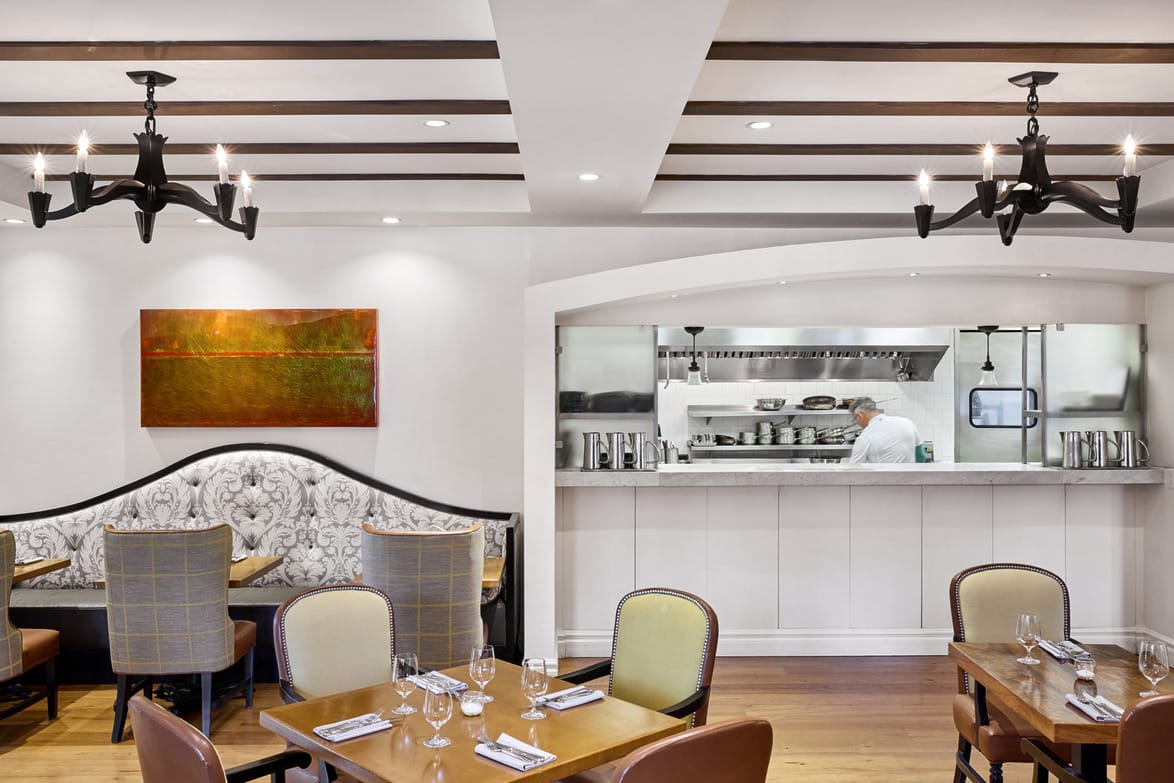 Elegant restaurant interior with cozy seating, wooden beams, chandeliers, and a view into the open kitchen with a chef cooking.