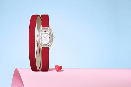 Red strap watch with diamond accents on pink and blue background, accompanied by a small red heart.