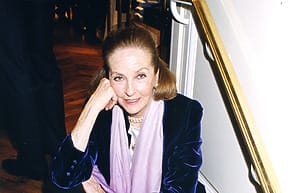Elderly woman sitting indoors, wearing a blue velvet jacket and a pink scarf, resting her chin on her hand.