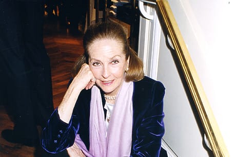 Elderly woman sitting indoors, wearing a blue velvet jacket and a pink scarf, resting her chin on her hand.