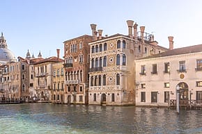 Historic buildings along the Grand Canal in Venice under a clear blue sky with calm waters in the foreground.