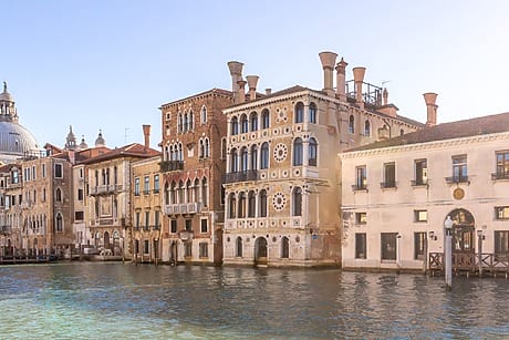 Historic buildings along the Grand Canal in Venice under a clear blue sky with calm waters in the foreground.