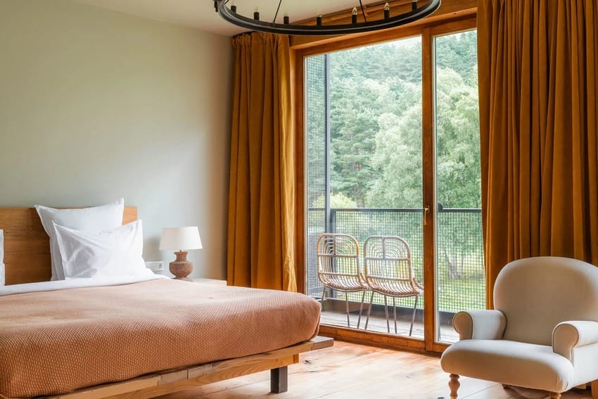 Cozy bedroom with a large bed, white pillows, orange curtains, armchair, and scenic window view of trees.
