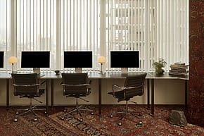 Modern office space with four computers on desks, ergonomic chairs, large windows, and patterned red carpet.