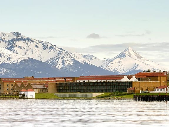 Historic prison building with snow-capped mountains in the background and calm water in the foreground.