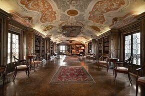 Ornate library with frescoed ceiling, vintage furniture, bookshelves lining the walls, and patterned carpets on wooden floor.