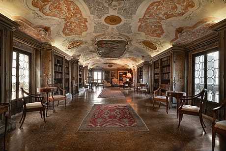 Ornate library with frescoed ceiling, vintage furniture, bookshelves lining the walls, and patterned carpets on wooden floor.
