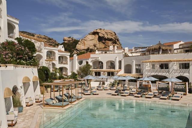 Mediterranean resort with white buildings, sun loungers, and a turquoise pool against a backdrop of rocky hills and blue sky