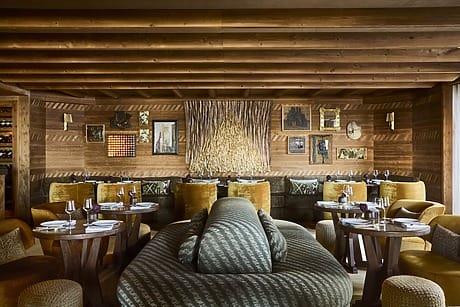 Cozy restaurant interior with wooden walls, green seating, round tables set for dining, and eclectic wall art.