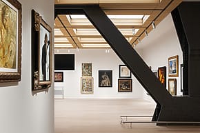 Modern art gallery interior with various framed paintings on white walls, featuring a large metal beam in the foreground.