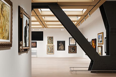Modern art gallery interior with various framed paintings on white walls, featuring a large metal beam in the foreground.
