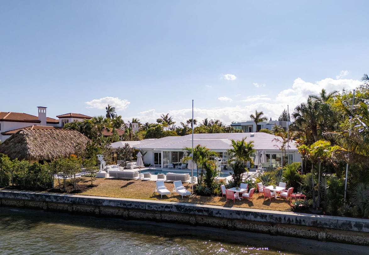 Scenic view of a waterfront property with a modern house, lush palm trees, and outdoor seating under a clear blue sky.