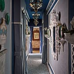 Elegant hallway with blue walls, ornate mirrors, decorative plates, and hanging lanterns, leading to a room with art on the walls.