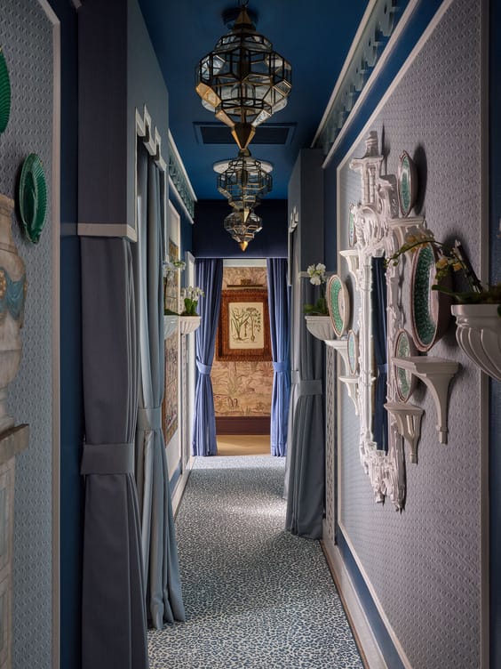 Elegant hallway with blue walls, ornate mirrors, decorative plates, and hanging lanterns, leading to a room with art on the walls.