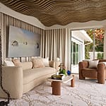 Modern living room with sculpted ceiling, beige sofa, brown armchair, glass door, abstract art on wall, and neutral decor.