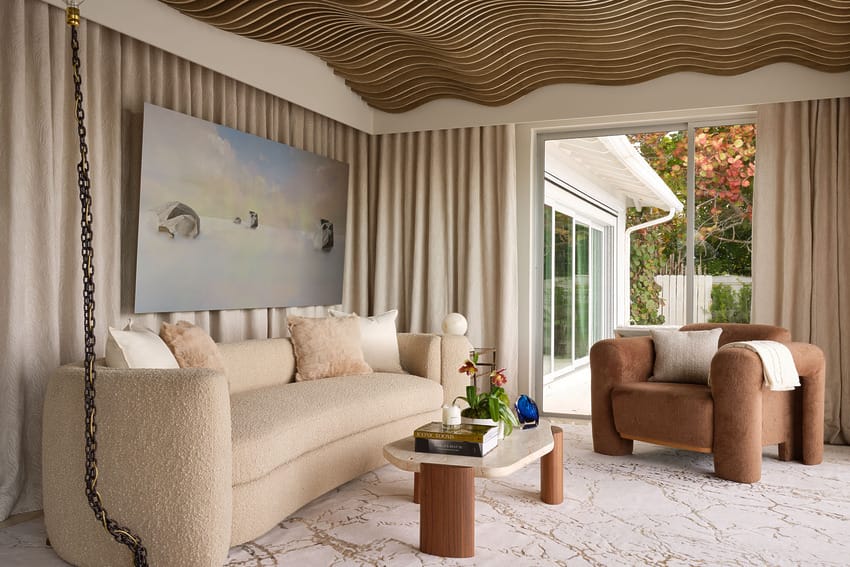 Modern living room with sculpted ceiling, beige sofa, brown armchair, glass door, abstract art on wall, and neutral decor.