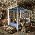 Ornate bedroom with floral wallpaper, canopy bed, patterned pillows, and matching drapes creating a cozy, vintage atmosphere.
