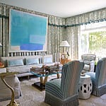 Elegant living room with pastel blue decor, large abstract painting, patterned curtains, and natural light from a window.