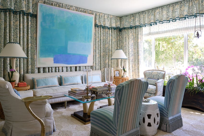 Elegant living room with pastel blue decor, large abstract painting, patterned curtains, and natural light from a window.