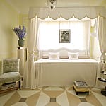 Elegant bedroom with a canopy bed, pale yellow walls, patterned floor, and decorative accents including framed photos and flowers.