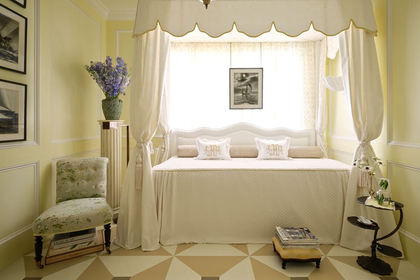 Elegant bedroom with a canopy bed, pale yellow walls, patterned floor, and decorative accents including framed photos and flowers.