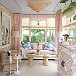 Bright, cozy sitting room with wicker furniture, floral wallpaper, large windows, pink curtains, and decorative ceiling light.