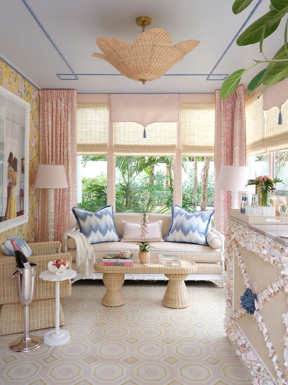 Bright, cozy sitting room with wicker furniture, floral wallpaper, large windows, pink curtains, and decorative ceiling light.