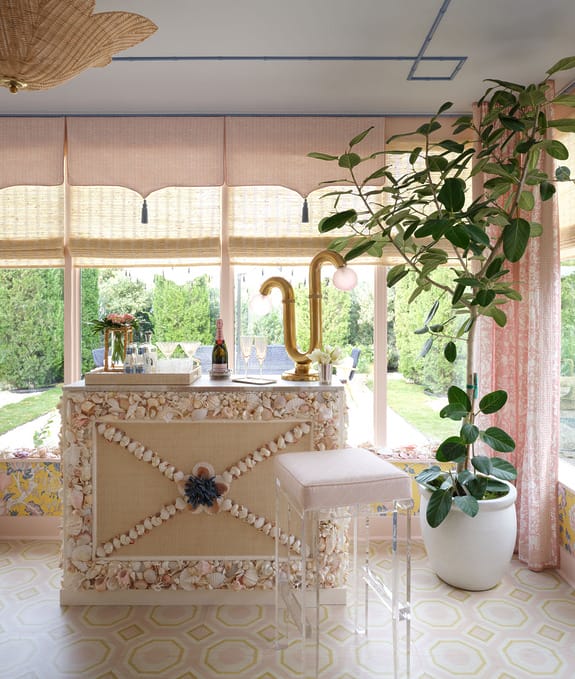 Indoor bar area with seashell decor, transparent stool, and large plant, set against a backdrop of large windows and greenery.