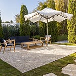 Outdoor seating area with chairs, a sofa, a coffee table, and a white umbrella on a gravel patio surrounded by greenery.