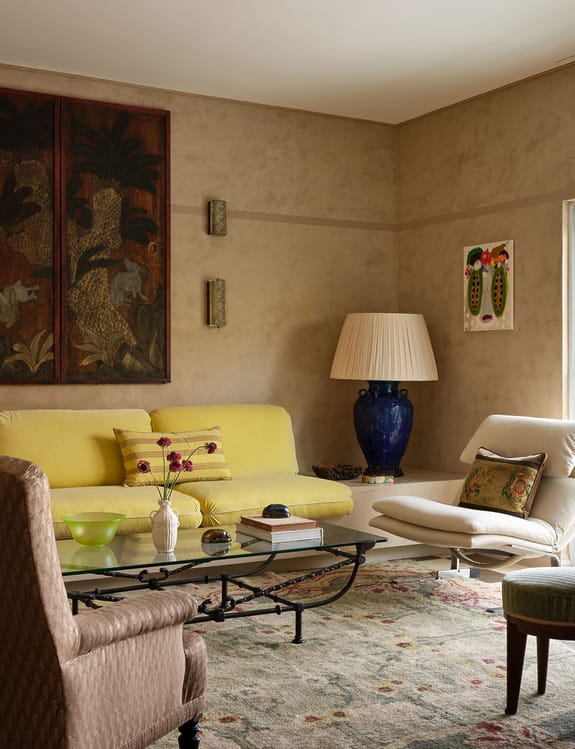 Cozy living room with yellow sofa, vintage armchairs, colorful rug, and elegant decorations including art and a large blue lamp.