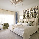 Modern bedroom with a plush bed, decorative pillows, curtains, and two green armchairs under a stylish ceiling light fixture.