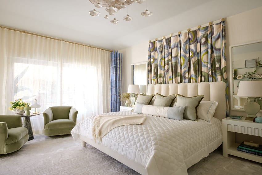 Modern bedroom with a plush bed, decorative pillows, curtains, and two green armchairs under a stylish ceiling light fixture.