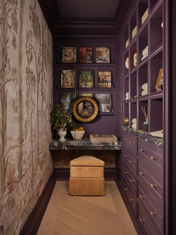 Elegant purple room with wall art, bookshelves, decorative items, and a stool, featuring rich textures and warm lighting.
