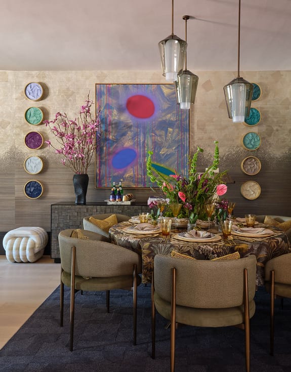 Modern dining room with a round table, vibrant artwork on wall, elegant pendant lights, and decorative plants.