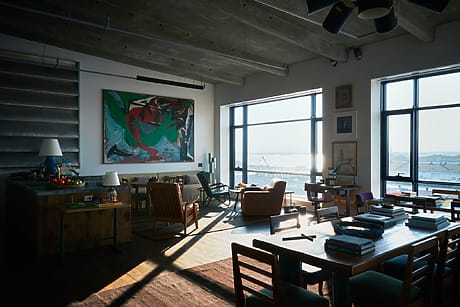 Spacious industrial-style loft with modern art, large windows, and warm afternoon sunlight streaming in.