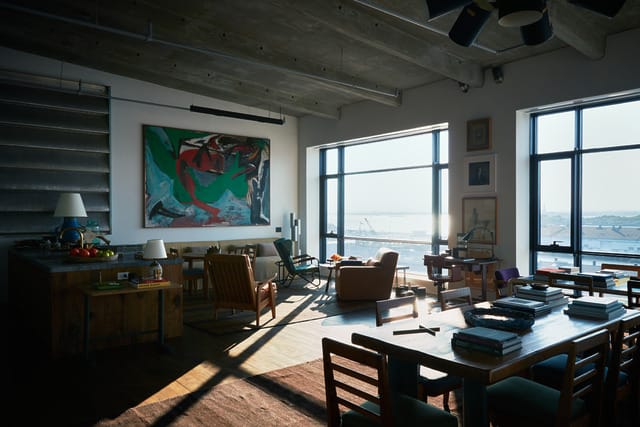 Spacious industrial-style loft with modern art, large windows, and warm afternoon sunlight streaming in.