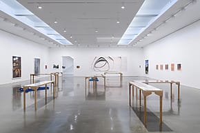 Modern art gallery interior with various framed artworks on white walls and minimalist tables showcasing displays.