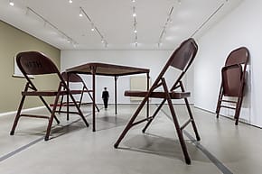 Oversized brown folding chairs and table art installation in contemporary gallery setting with a visitor in the background.