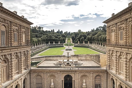 View of the Boboli Gardens in Florence, a grand architectural courtyard with a fountain and lush green gardens under a partly cloudy sky.