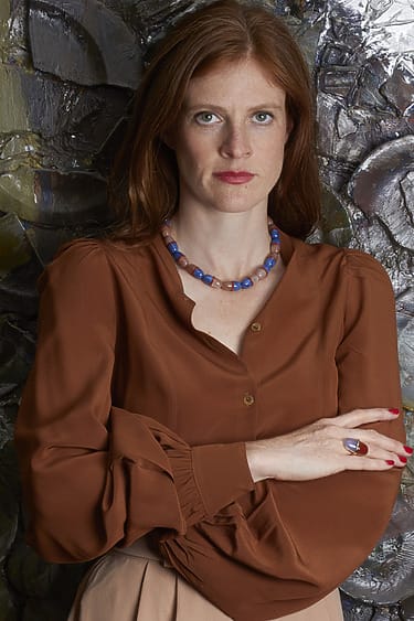 Woman with red hair wearing a brown blouse and necklace, standing against an abstract textured background.