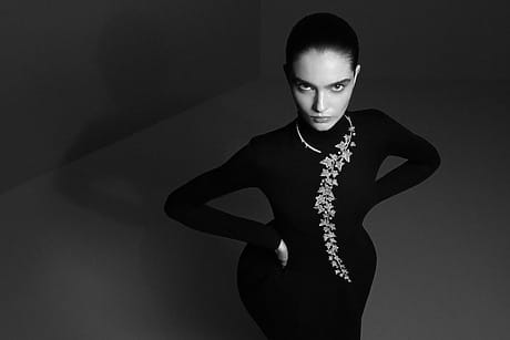 Person in elegant black dress with decorative floral design, standing confidently in a minimalist gray background.