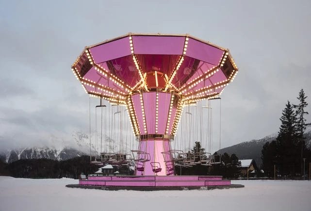 Pink illuminated swing carousel in a snowy winter landscape with trees and cloudy sky in the background.