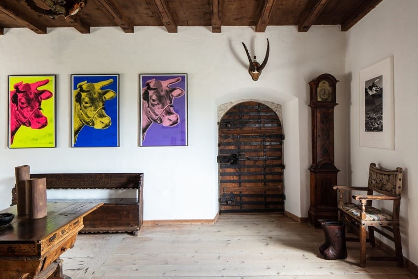 Rustic room with wooden ceiling, vibrant cow artwork, vintage furniture, and an antique clock by an old wooden door.
