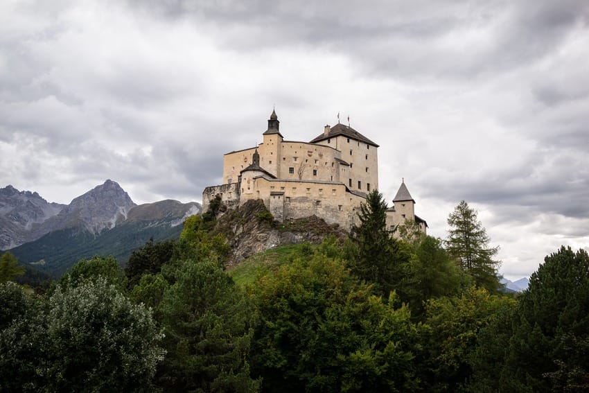 Medieval castle on a rocky hill surrounded by dense forest and mountains under a cloudy sky.